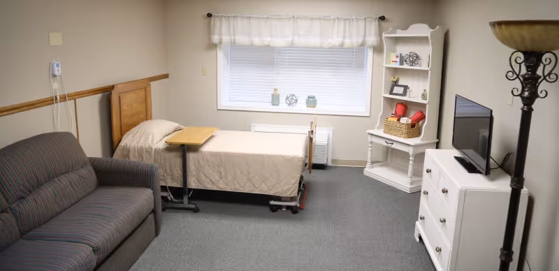 A small, neatly arranged room with a single bed covered in a beige quilt, a striped couch, a white dresser with a flat-screen TV on top, a white corner shelf with decorative items, and a tall floor lamp. A window with white blinds and a sheer curtain is centered on the back wall.
