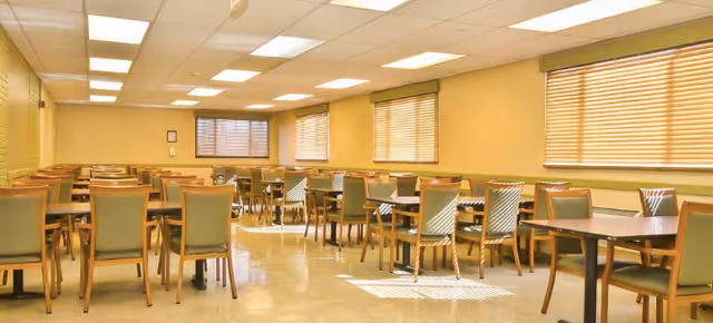 A spacious dining room with multiple wooden tables and green cushioned chairs arranged neatly. The room has large windows with blinds allowing natural light to fill the space, and a ceiling with recessed lighting panels.