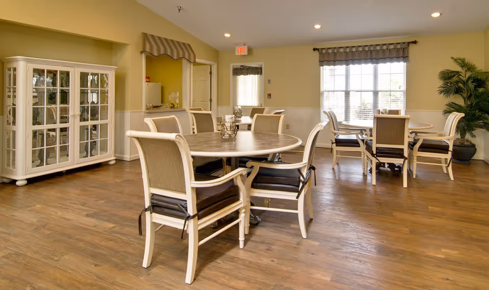 A bright and spacious dining room in St. Francis Park Senior Living with round tables and cushioned chairs arranged neatly on a wooden floor. The room features large windows with curtains allowing natural light to fill the space, a white display cabinet, and a potted plant in the corner.