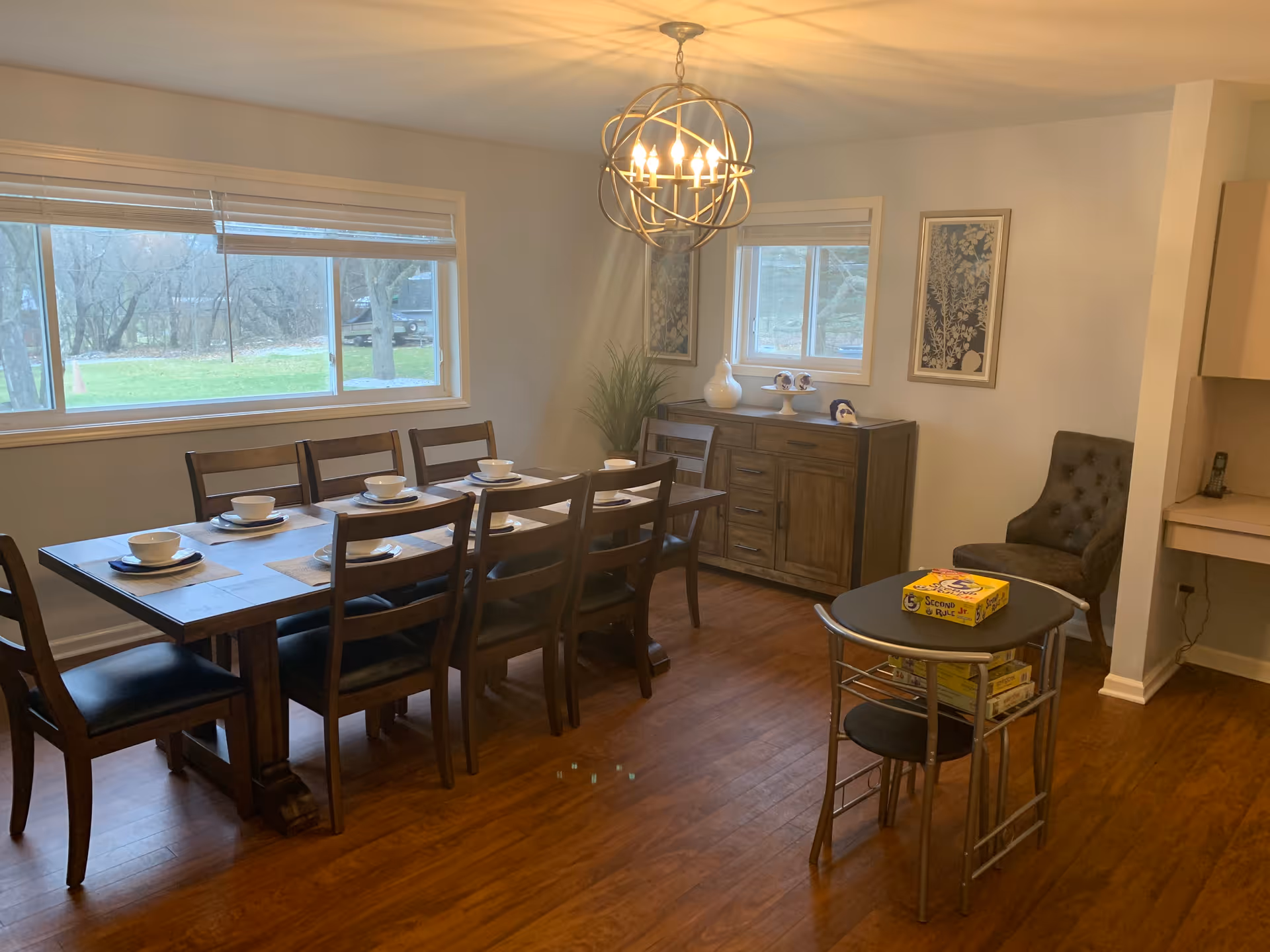 A dining room with a large wooden dining table set with eight chairs, each place set with a bowl and plate. The room has two windows with views of trees and grass outside. There is a wooden sideboard against the wall with decorative items and framed artwork above it. A modern chandelier hangs from the ceiling. In the corner, there is a small table with board games on top and a cushioned chair nearby. The floor is wooden.