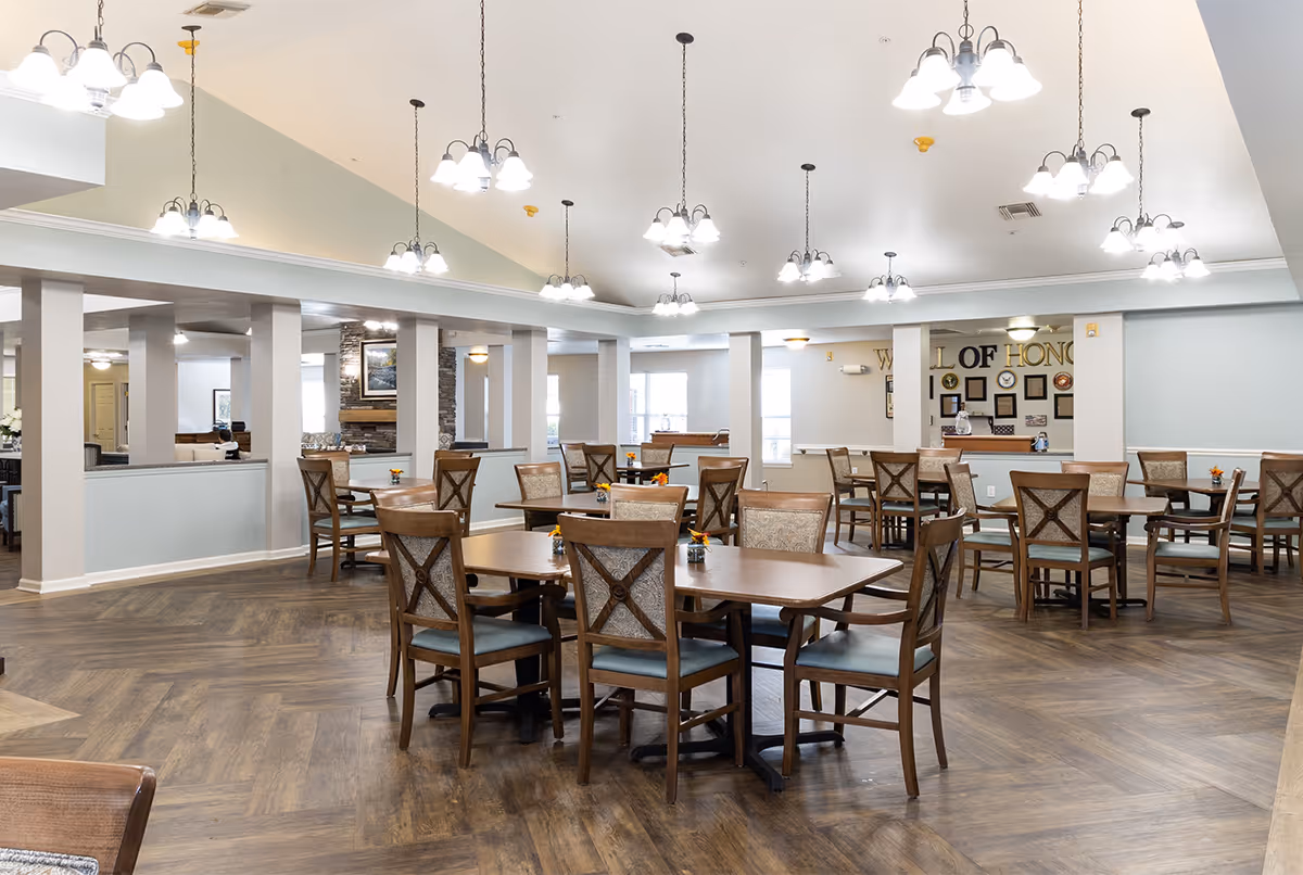 A spacious dining room with multiple wooden tables and chairs arranged neatly. The room features a high ceiling with several hanging light fixtures, wood-patterned flooring, and a fireplace in the background. The walls are light-colored, and there is a 'Wall of Honor' display on one side.