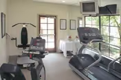 Small fitness room with exercise equipment including a treadmill, stationary bike, and weight machine. The room has large windows letting in natural light and a wall-mounted television in the corner. There is a door at the back and a small table with a white cloth near the windows.