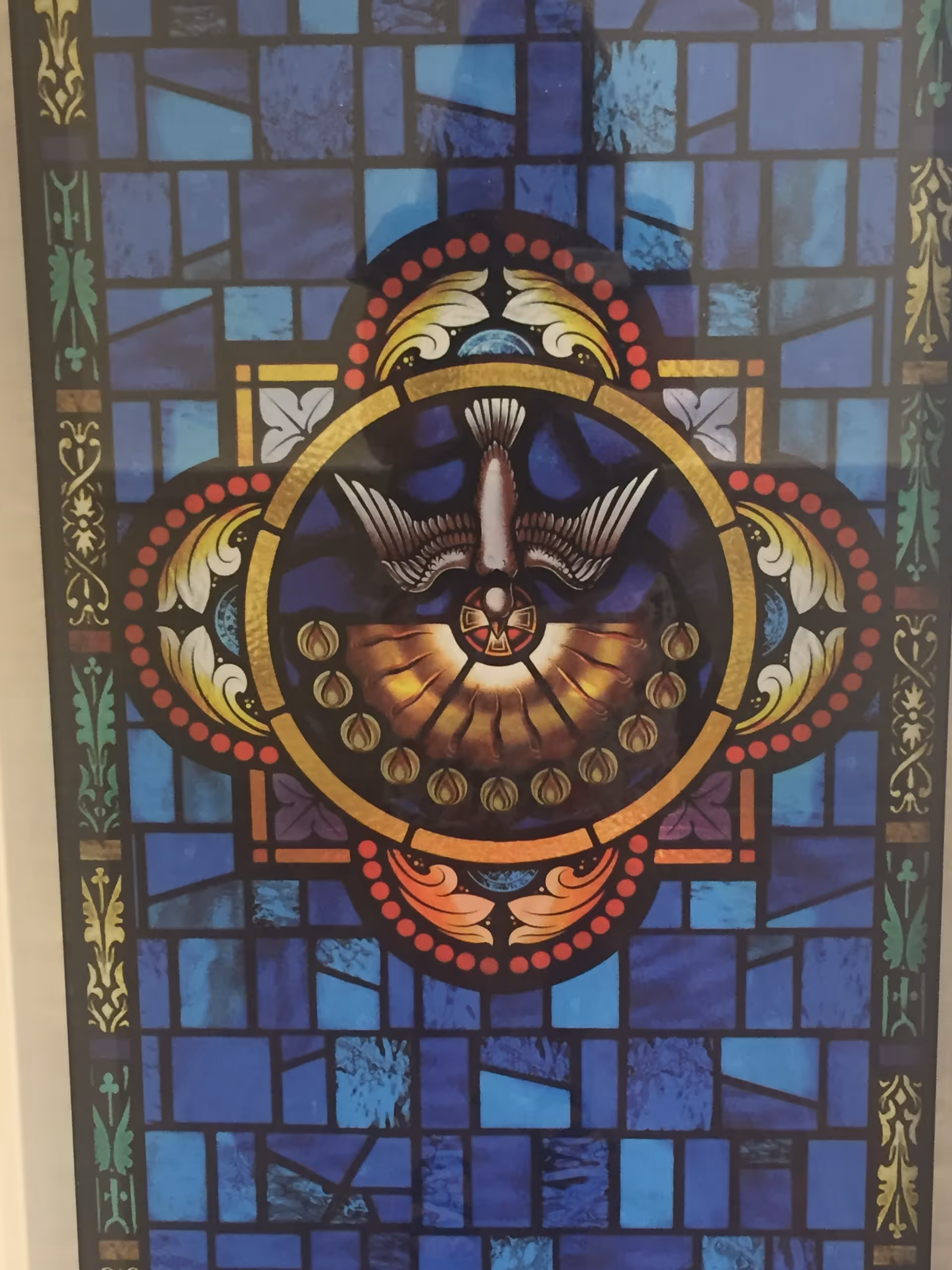 A stained glass window featuring a central circular design with a dove in flight surrounded by decorative patterns and a background of blue geometric shapes.