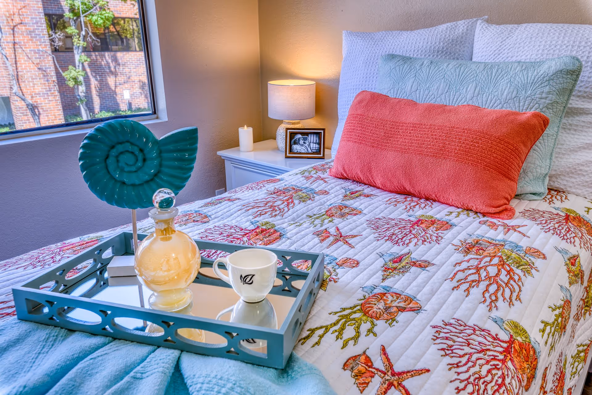 A neatly made bed with a colorful sea-themed quilt featuring shells and coral, accented with a large coral-colored pillow and a light blue pillow. On the bed is a decorative tray holding a glass bottle, a white cup with a leaf design, and a teal spiral shell ornament. A bedside table with a lit lamp, a candle, and a framed black and white photo is visible next to the bed. A window shows a view of a brick building and some trees outside.