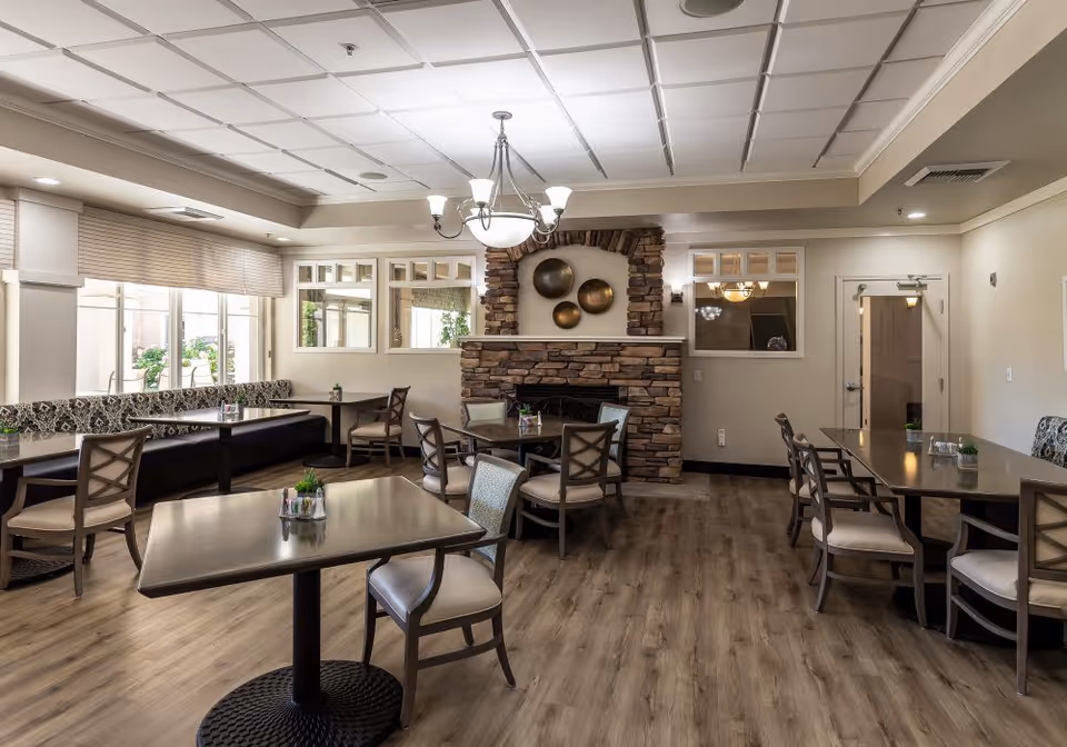 Communal dining room with tables and chairs, a stone fireplace, chandelier, and large windows.