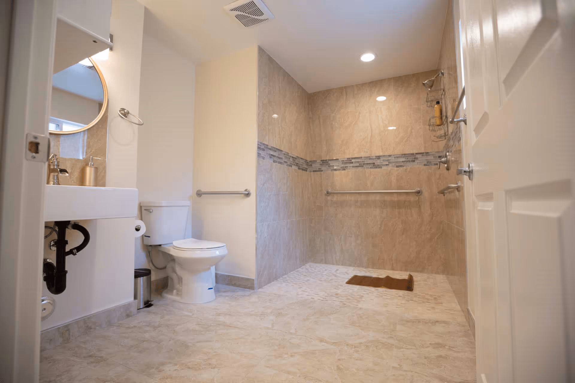 A clean, spacious bathroom with beige tiled floors and walls. The bathroom features a white toilet, a white sink with a gold soap dispenser, a round mirror with a gold frame, and a walk-in shower area with grab bars and a small brown mat on the floor.