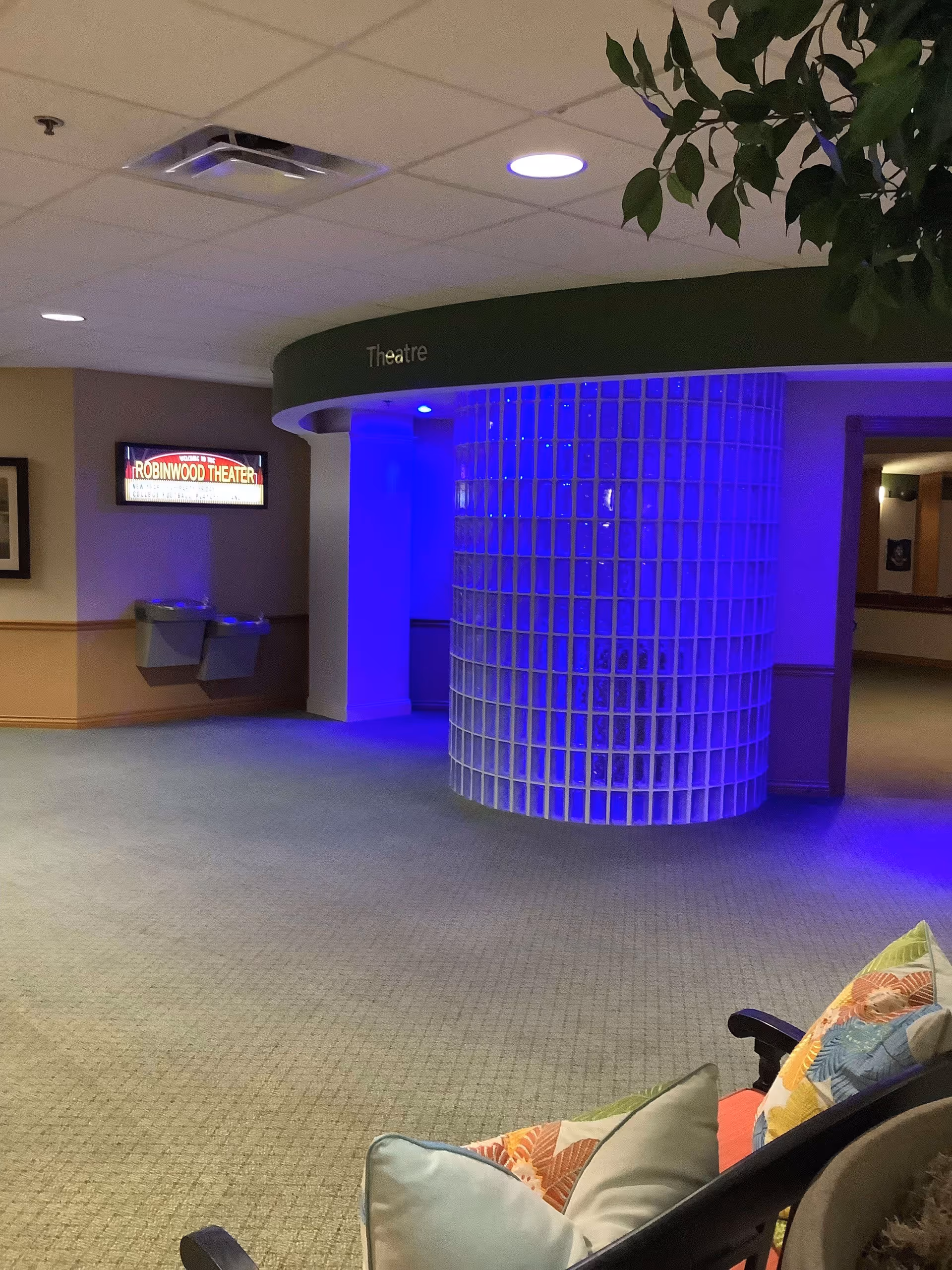 Interior hallway area of Robinwood Retirement Resort featuring a curved glass block wall illuminated with blue lighting. A sign above the entrance reads 'Theatre'. To the left, a lighted sign says 'Robinwood Theater'. There are two water fountains mounted on the wall beneath the sign. In the foreground, a bench with colorful pillows is partially visible. The ceiling has recessed lighting and an air vent.