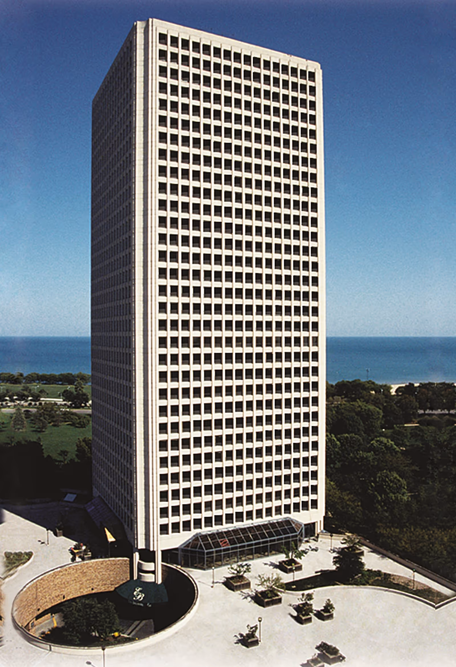 Tall rectangular high-rise building with many windows, surrounded by trees and a paved area with planters, with a body of water visible in the background under a clear blue sky.