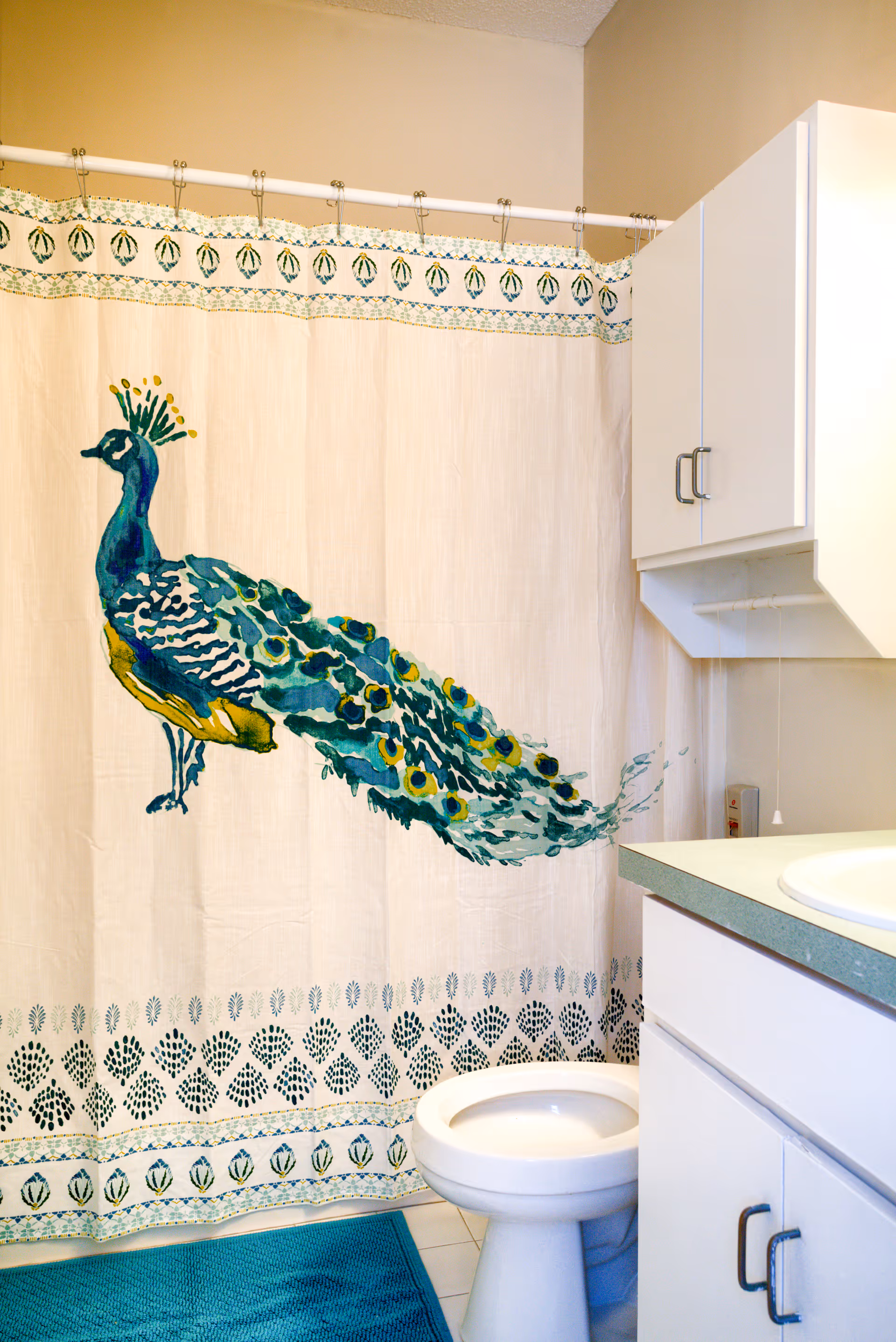Bathroom with a white toilet, white cabinets, and a shower curtain featuring a large colorful peacock design. There is a blue bath mat on the floor.
