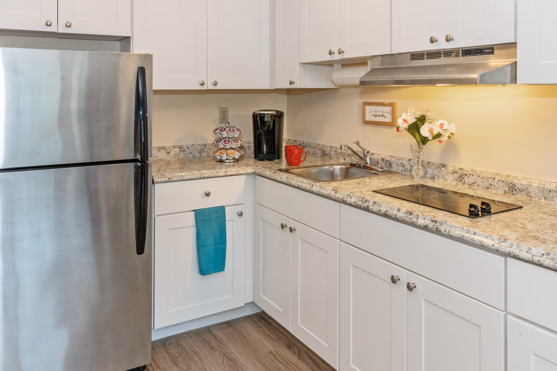 A modern kitchen with white cabinets, a stainless steel refrigerator, a granite countertop, a built-in electric cooktop, a stainless steel sink, a coffee maker, a red mug, a small vase with white flowers, and a blue towel hanging on a cabinet door.