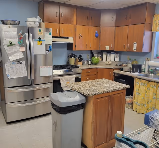 Communal kitchen with a stainless steel French-door refrigerator, gas range, wooden cabinets, a small granite-topped island, and a trash can.
