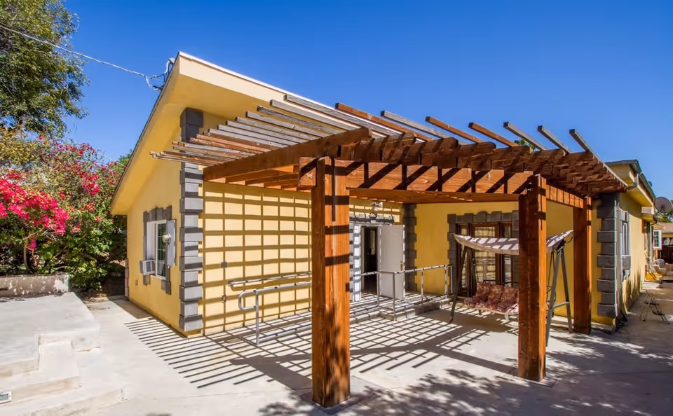 Outdoor patio area at Bentley Hills by Serenity Care Health featuring a wooden pergola casting shadows on the concrete ground, a swing with cushions, yellow building walls with gray trim, and surrounding greenery including a flowering bush.