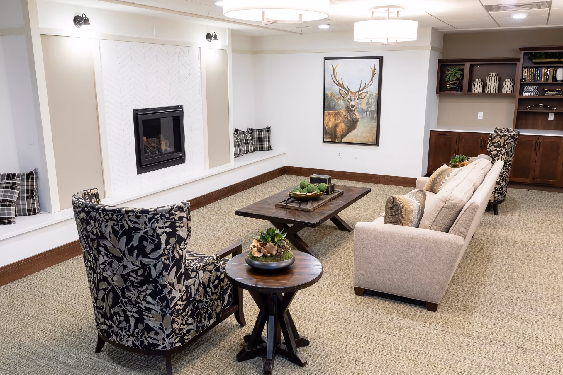 A cozy living room area in a senior living facility featuring a beige sofa with cushions, two patterned armchairs, a wooden coffee table with decorative items, a small round side table with a plant, a built-in fireplace with plaid pillows on the bench seating, a large painting of a deer on the wall, and built-in wooden shelves and cabinets in the background.