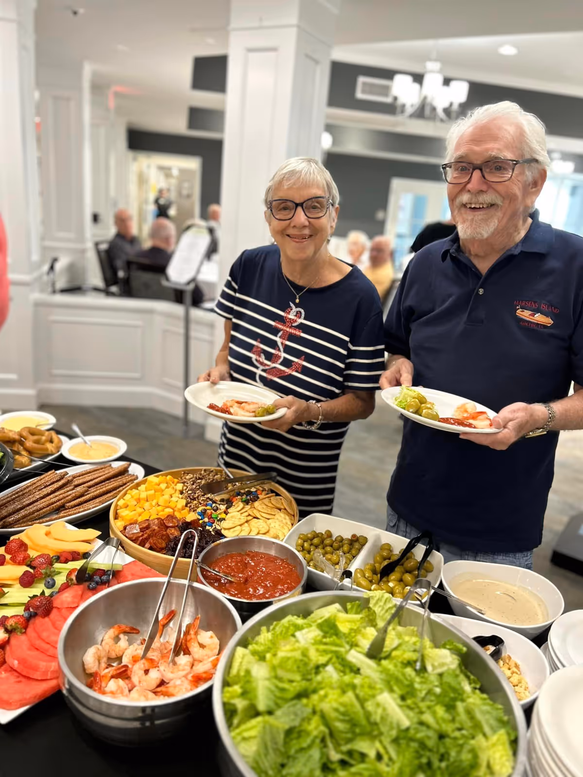 Two smiling senior residents serve themselves from a buffet table with salad, shrimp, fruit and snacks in a dining room.