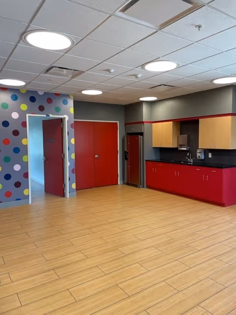 Spacious interior common room with wood-look flooring, a kitchenette with red cabinets, and a polka-dot accent wall with red doors.