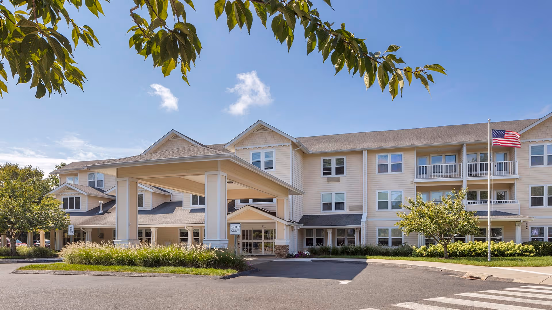 Exterior view of a three-story senior living facility building with beige siding and multiple windows. There is a covered entrance with white pillars and a driveway in front. An American flag is flying on a flagpole near the entrance. Trees and landscaping with green grass and bushes surround the building under a clear blue sky.