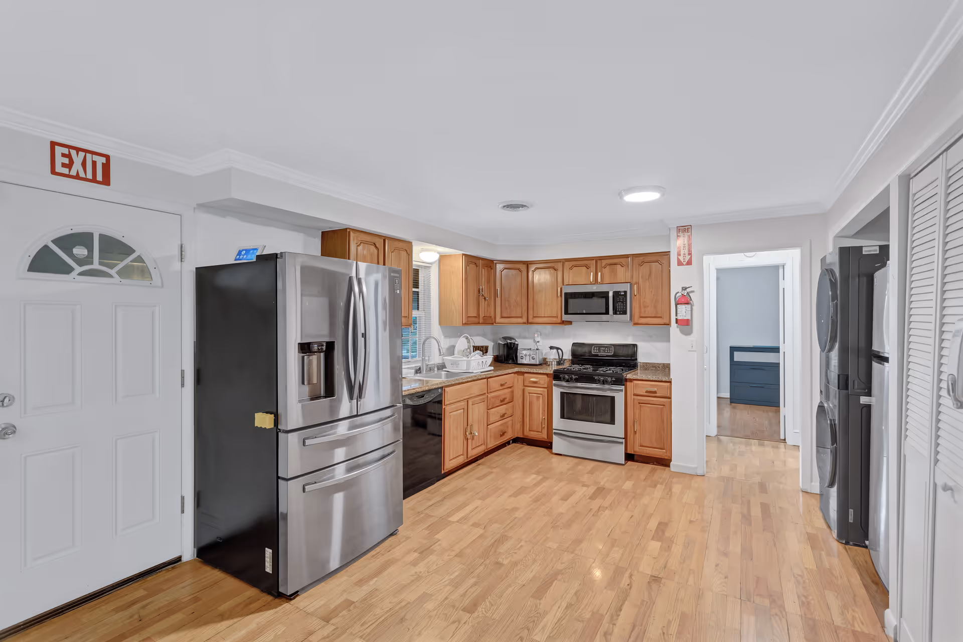 A clean and modern kitchen with wooden cabinets, stainless steel refrigerator, stove, microwave, and dishwasher. The kitchen has light hardwood floors and white walls. There is an exit door with a red exit sign above it on the left side. A fire extinguisher is mounted on the wall near a doorway leading to another room.
