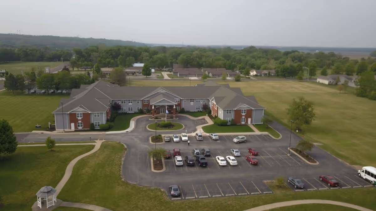 Aerial view of the John M. Evans Supportive Living Community building surrounded by green lawns and trees, with a parking lot in front containing several parked cars and a small white gazebo on the lawn.