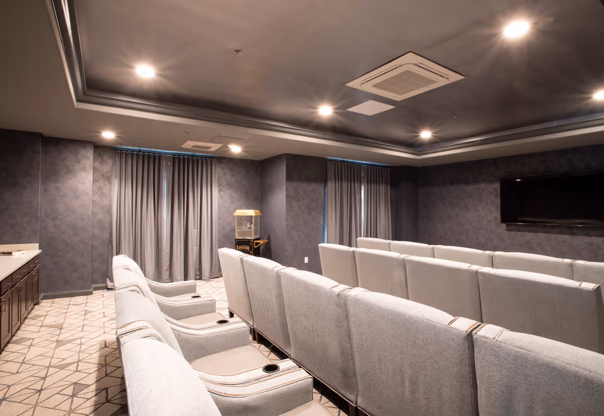 A small theater room with multiple rows of light gray upholstered seats facing a large flat-screen TV mounted on a dark wall. The room has a patterned carpet, dark walls, ceiling lights, and a popcorn machine in the corner near closed curtains.