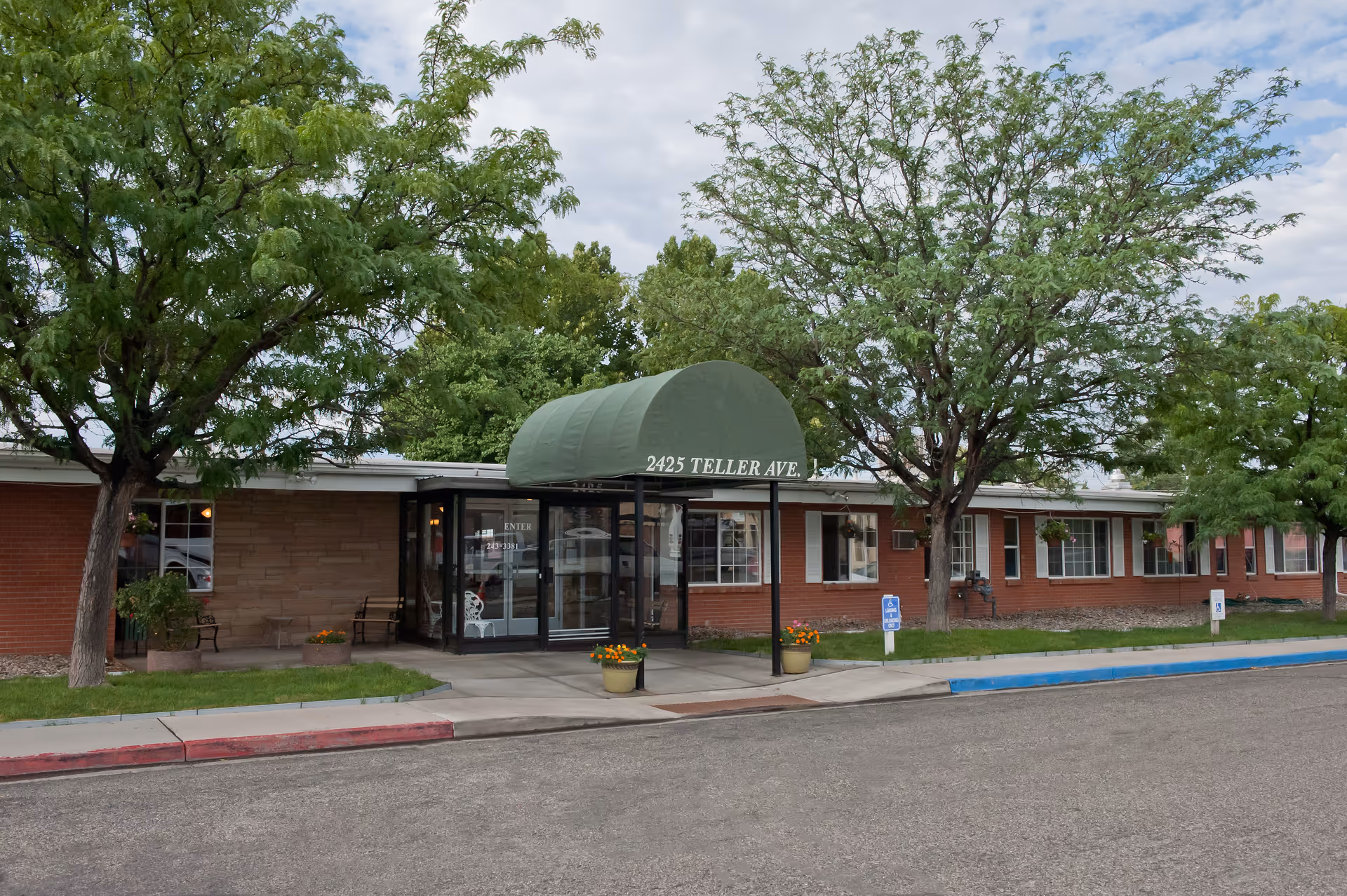 Exterior view of a single-story brick building with a green awning over the entrance that reads '2425 TELLER AVE.'. There are trees and potted plants near the entrance, and a sidewalk leading to the door. The sky is partly cloudy.