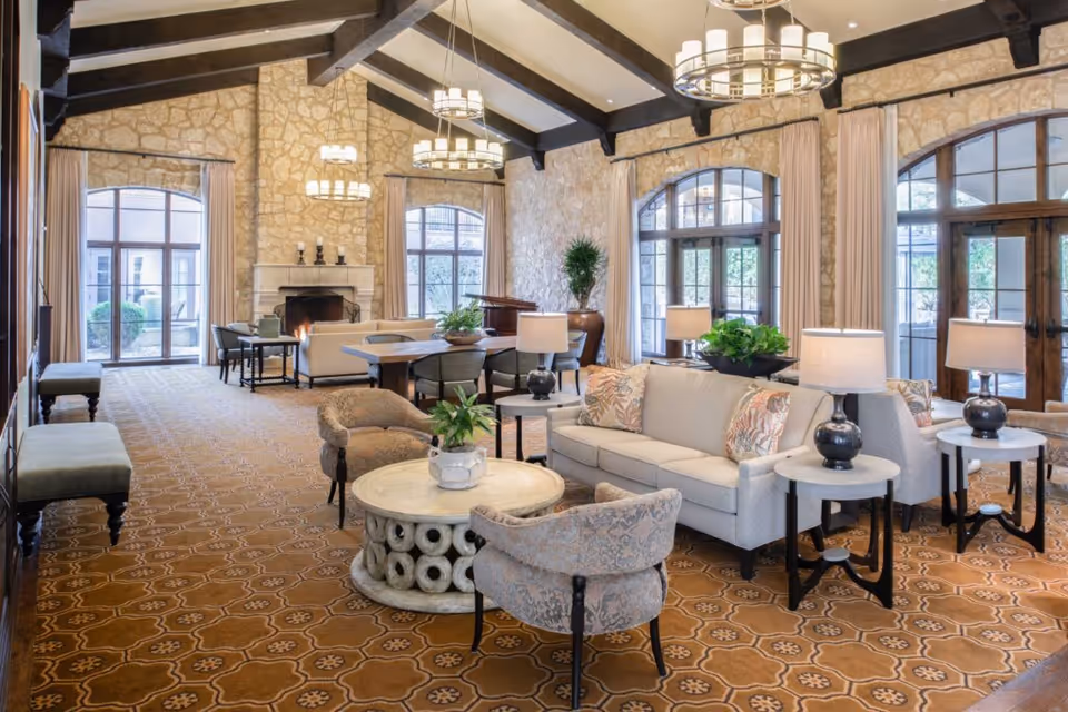 Spacious common lounge with sofas, armchairs, round tables, chandeliers, a fireplace, and large arched windows.