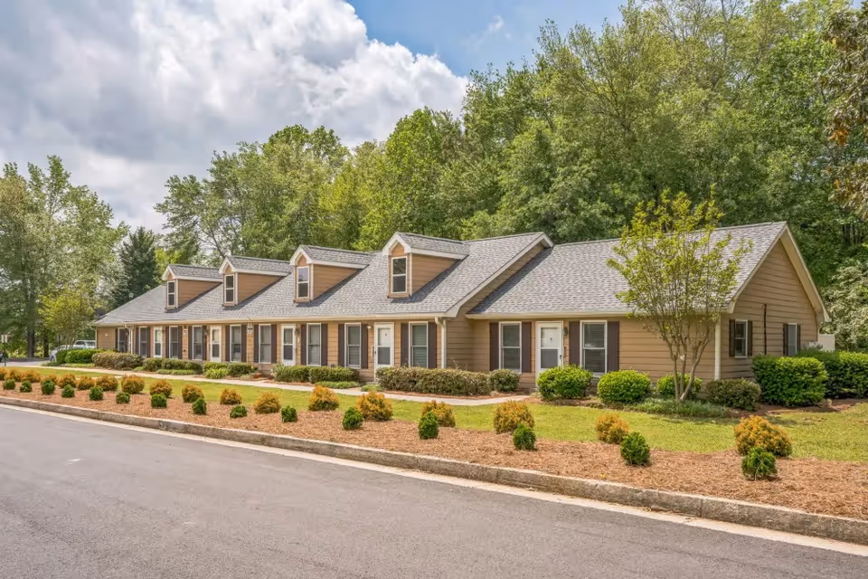 Exterior view of a single-story residential building with multiple entrances and dormer windows on the roof, surrounded by landscaped bushes and trees under a partly cloudy sky.