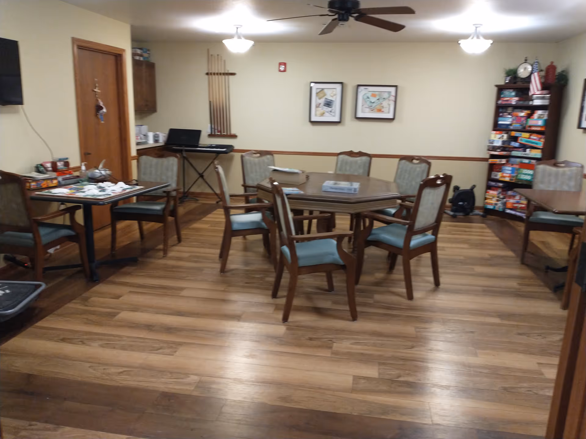 A common room with wooden flooring and several tables and chairs arranged for group activities. One table has board games and puzzles on it. There is a keyboard against the wall, a rack with pool cues, framed pictures on the wall, and a bookshelf filled with board games and other items. The room is lit by ceiling lights and a ceiling fan.