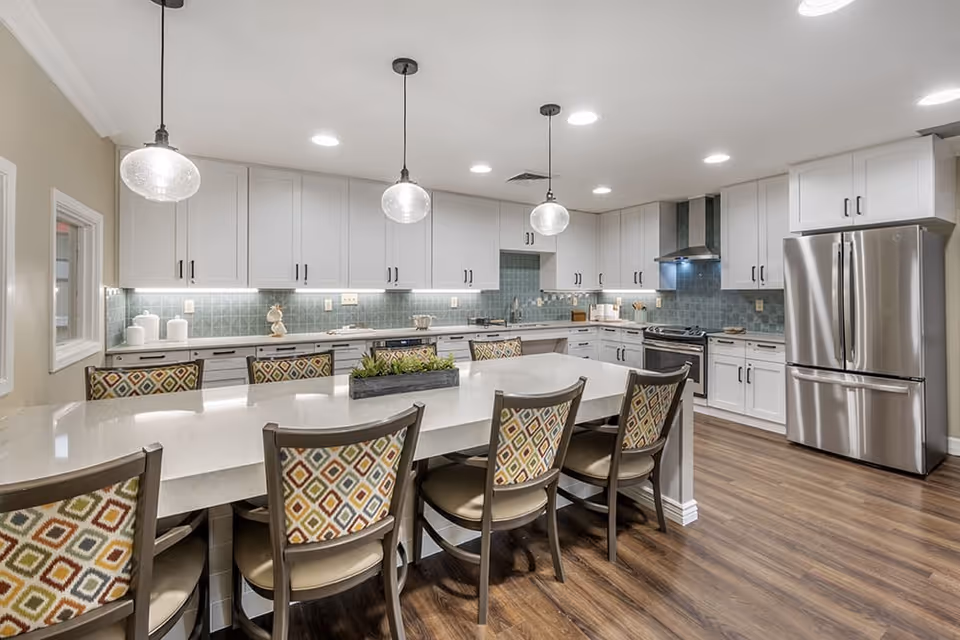 Modern communal kitchen and dining area with a large island, patterned chairs, white cabinetry, stainless-steel appliances, and pendant lights.