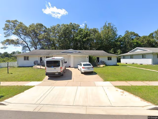 Single-story white ranch-style building with two vehicles parked in the driveway, a front lawn, and trees in the background under a clear blue sky.