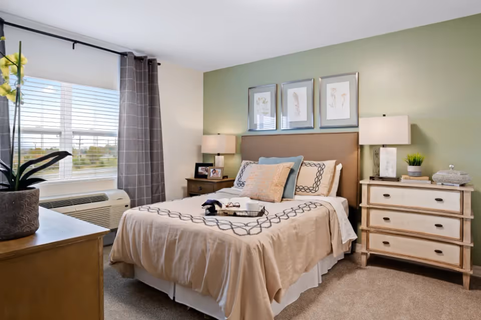 A cozy bedroom with a neatly made bed featuring beige and patterned bedding. There are two bedside tables with lamps, framed botanical artwork above the headboard, a window with gray curtains, and a potted plant on a wooden dresser.
