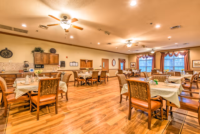 A spacious dining room in an assisted living facility with multiple tables covered with white tablecloths and set with glassware and small flower arrangements. The room features wooden flooring, ceiling fans with lights, large windows with decorative valances, and wooden chairs with cushioned seats. The walls are decorated with clocks, framed pictures, and cabinets.