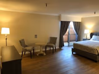 A spacious bedroom with a large bed, two chairs and a small round table near a sliding glass door with curtains leading to a balcony. The room has wooden flooring, a floor lamp, and a dresser against one wall.