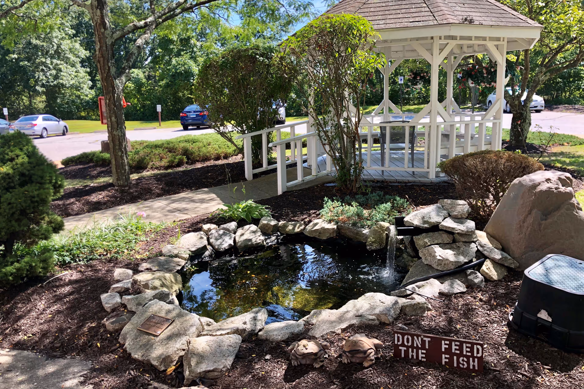 A small outdoor garden area featuring a white wooden gazebo with a ramp, surrounded by trees and shrubs. In the foreground, there is a small pond with rocks around it and a sign that reads 'DON'T FEED THE FISH'. Several cars are parked in the background along a paved road.