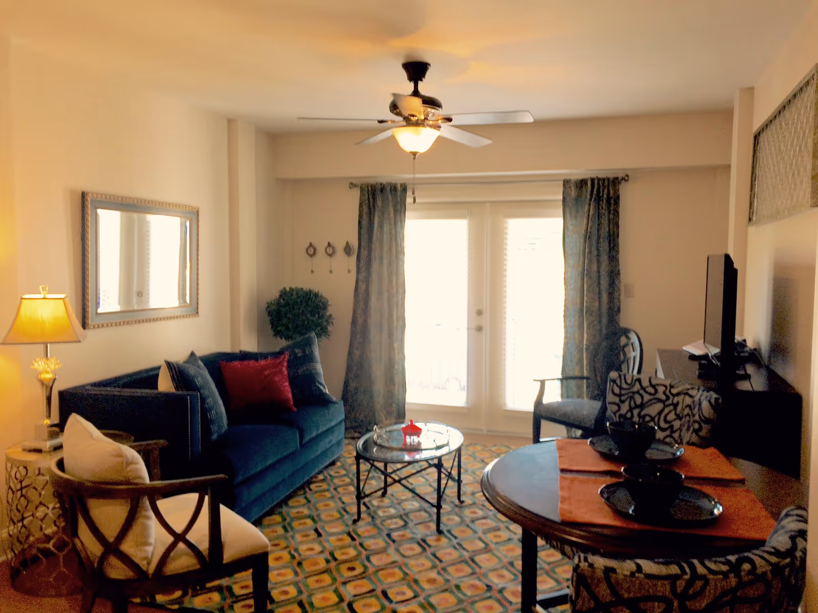 A cozy living room with a blue sofa adorned with pillows, a round glass coffee table, a patterned carpet, a ceiling fan with light, a lamp on a side table, two chairs around a round dining table set with black plates and orange placemats, and large windows with curtains letting in natural light.