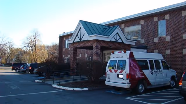 Front entrance of a brick senior living building with a covered portico and a wheelchair-accessible transport van parked in the lot.