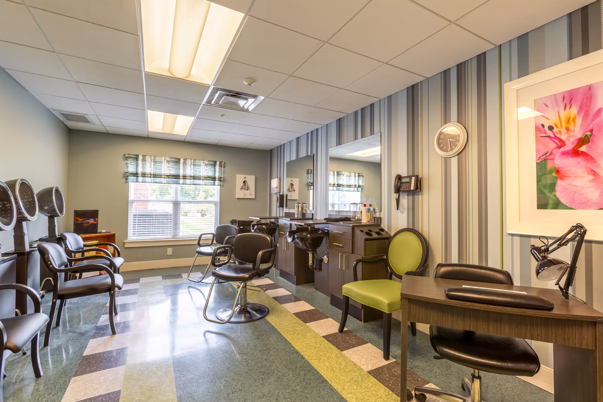 Interior view of a hair salon area in a senior living facility with several black salon chairs, hair dryers, mirrors, and a small desk with a green chair and a black office chair. The room has striped wallpaper, a clock, a framed flower picture, and a window with a patterned valance.
