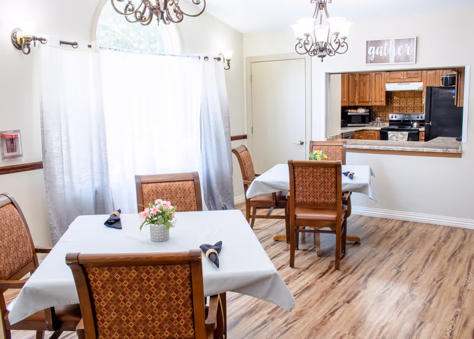 A bright dining room with two tables covered in white tablecloths, each surrounded by four wooden chairs with patterned upholstery. A small flower arrangement is placed on one table. The room features light-colored walls, wood flooring, two chandeliers, and a large window with sheer curtains. A pass-through window reveals a kitchen with wooden cabinets and appliances. A decorative sign above the pass-through window reads 'gather'.