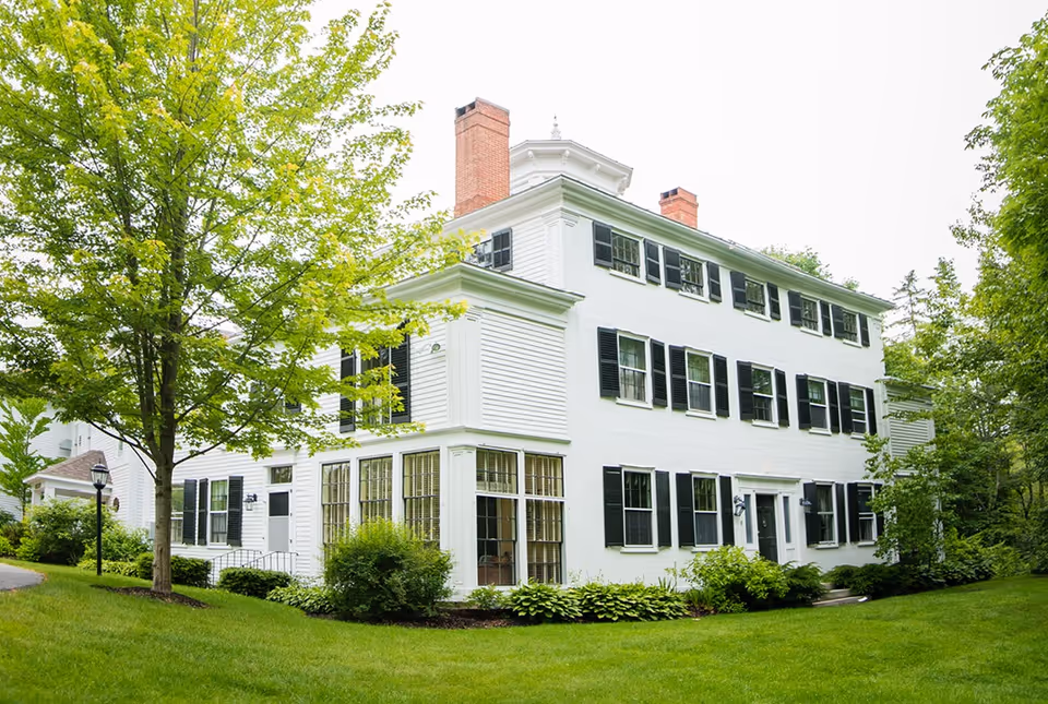 A large white multi-story building with black shutters and two brick chimneys, surrounded by green grass, bushes, and trees under a bright sky.