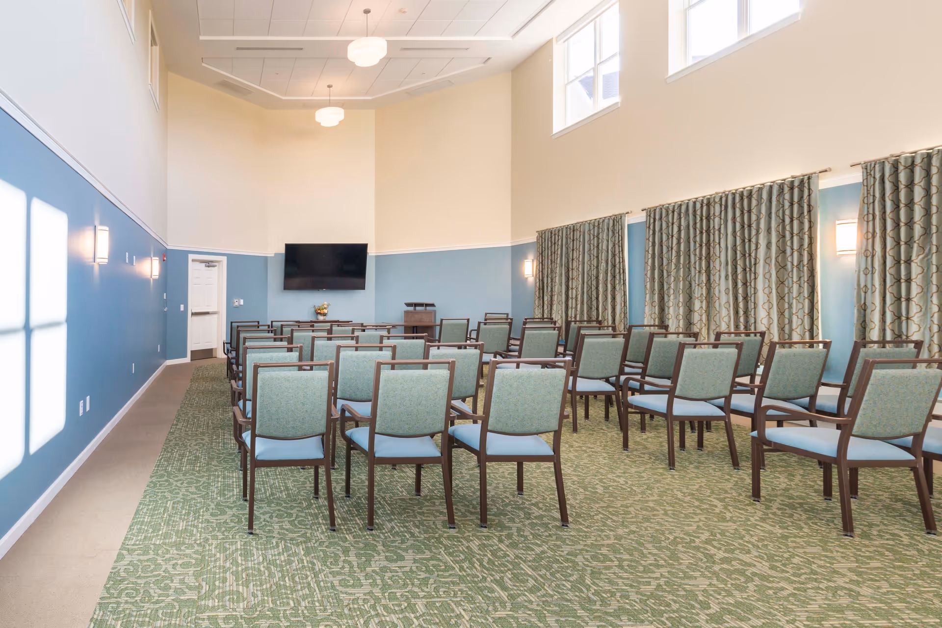 A conference or meeting room with rows of green cushioned chairs facing a wall-mounted flat screen TV. The room has high ceilings, blue and cream walls, patterned green carpet, and large windows near the ceiling letting in natural light. There are patterned curtains along one wall and wall-mounted lights.