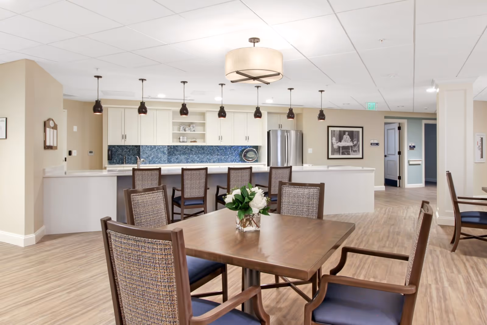 A modern senior living facility dining area with a wooden table and chairs in the foreground, a kitchen counter with bar stools, pendant lights hanging above, white cabinets, a stainless steel refrigerator, and a blue tile backsplash. The room has light-colored walls and wood flooring.