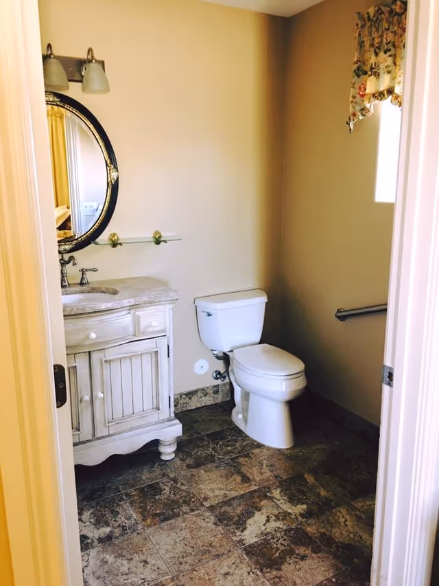 A small bathroom with a white toilet and a white wooden vanity with a marble countertop. Above the vanity is a round mirror with a decorative frame and two wall-mounted light fixtures. The floor is covered with brown and beige tiles, and there is a small window with a floral curtain on the right wall. A metal grab bar is installed next to the toilet.
