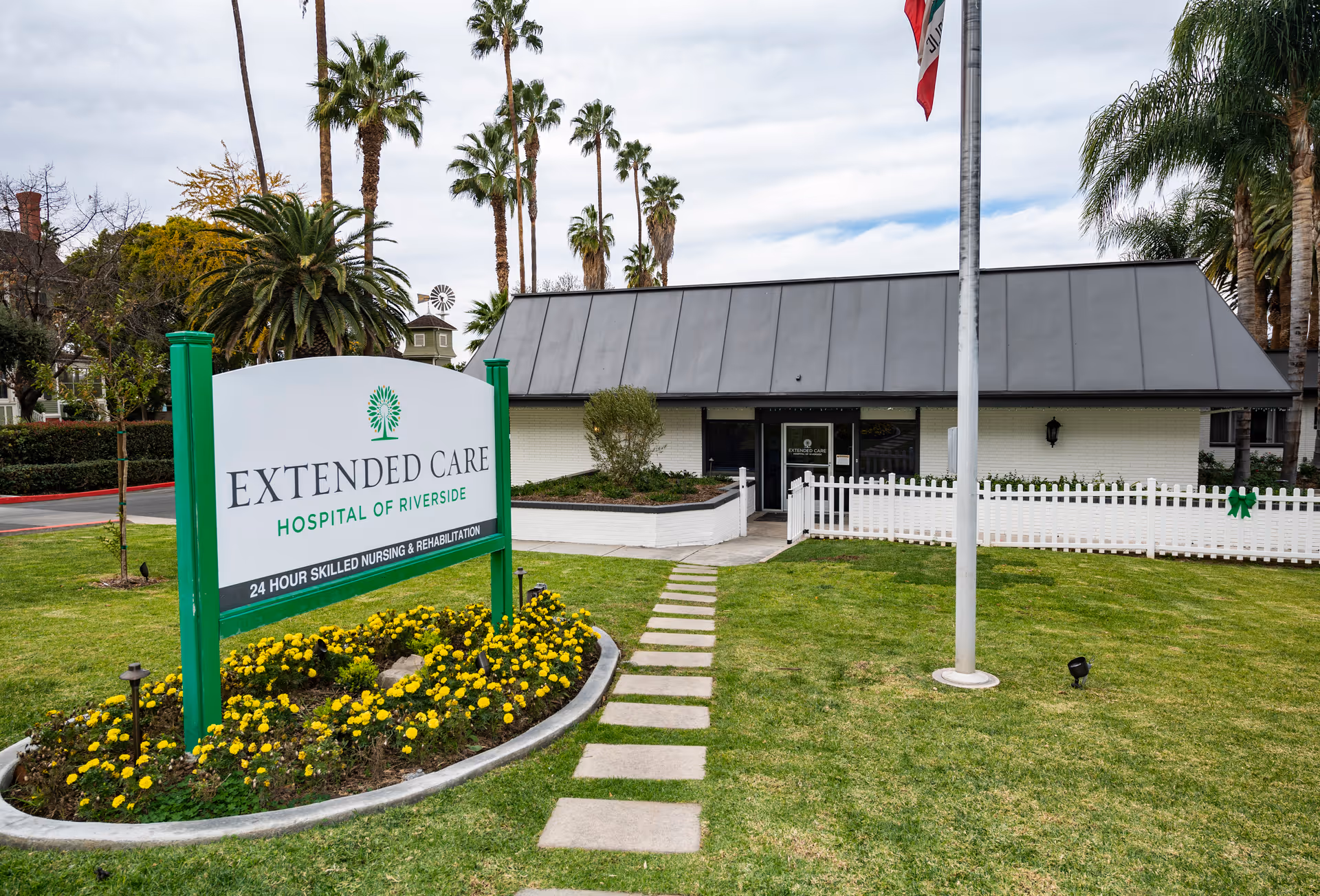 Exterior view of the Extended Care Hospital of Riverside building with a green lawn, a pathway leading to the entrance, palm trees in the background, and a sign in the foreground that reads 'Extended Care Hospital of Riverside 24 Hour Skilled Nursing & Rehabilitation'.