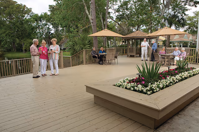 A spacious outdoor wooden deck area with several groups of elderly people socializing and sitting at tables under large beige umbrellas. The deck is surrounded by trees and greenery, with a raised planter filled with colorful flowers in the foreground.