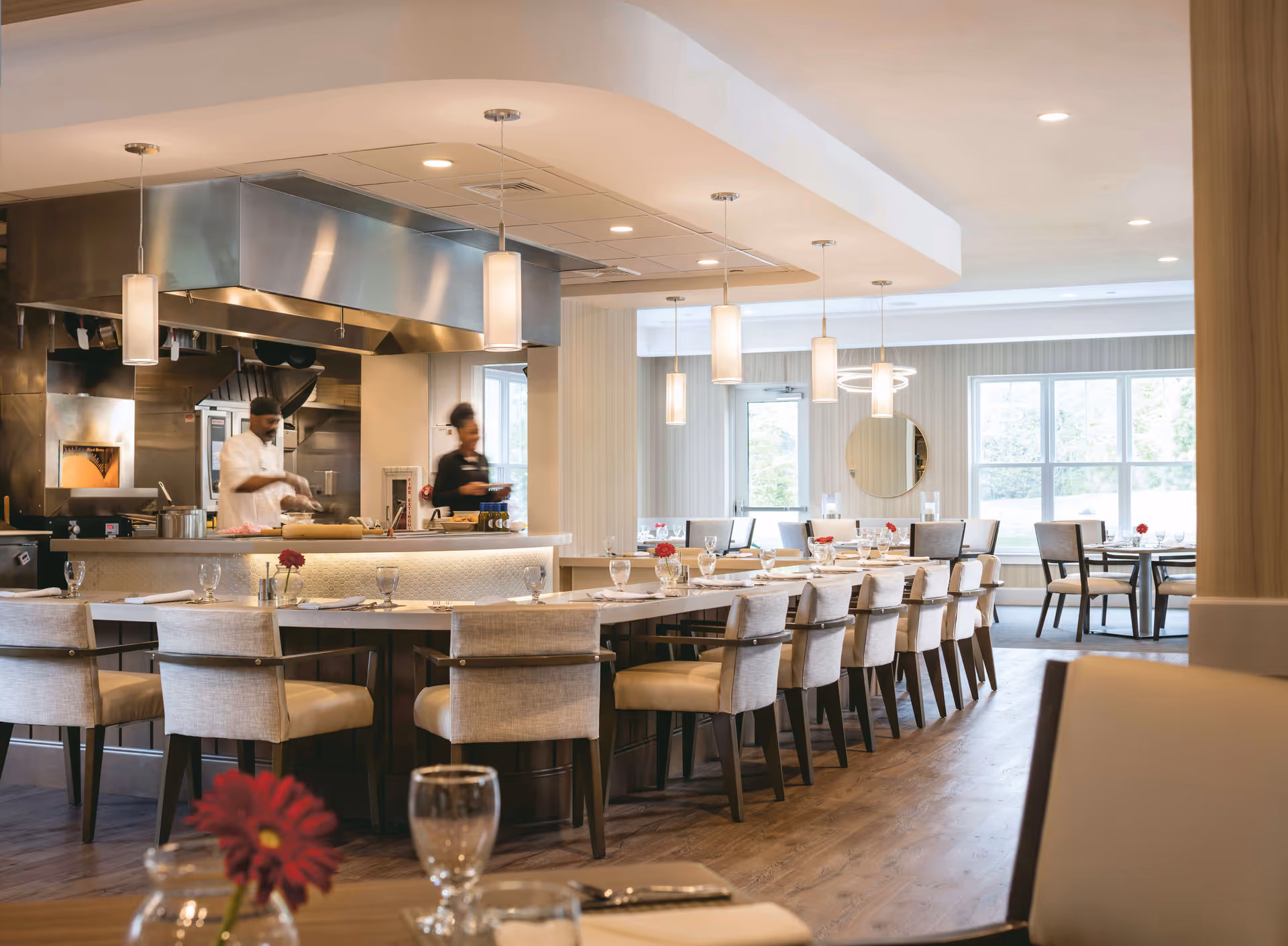 A modern dining area in a senior living facility with a long table surrounded by cushioned chairs, set with glasses, napkins, and red flowers in small vases. In the background, two staff members are working in an open kitchen with stainless steel appliances and hanging pendant lights.