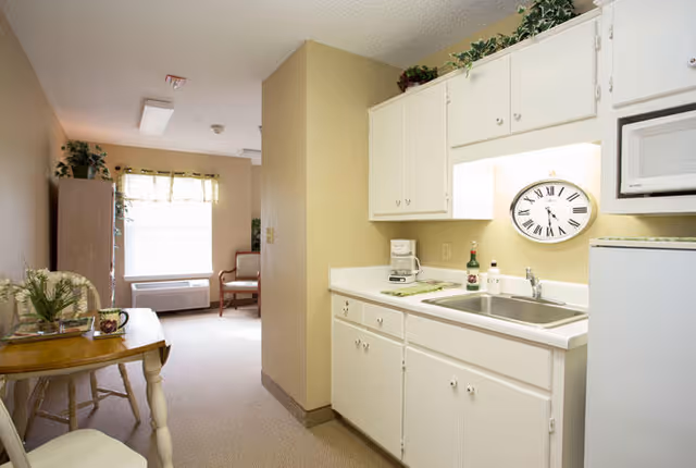 A small kitchenette area with white cabinets, a sink, a coffee maker, a microwave, and a refrigerator. A large clock is mounted on the wall above the sink. To the left, there is a small table with a plant and a cup, and further back, a window with a curtain and a chair near an air conditioning unit.