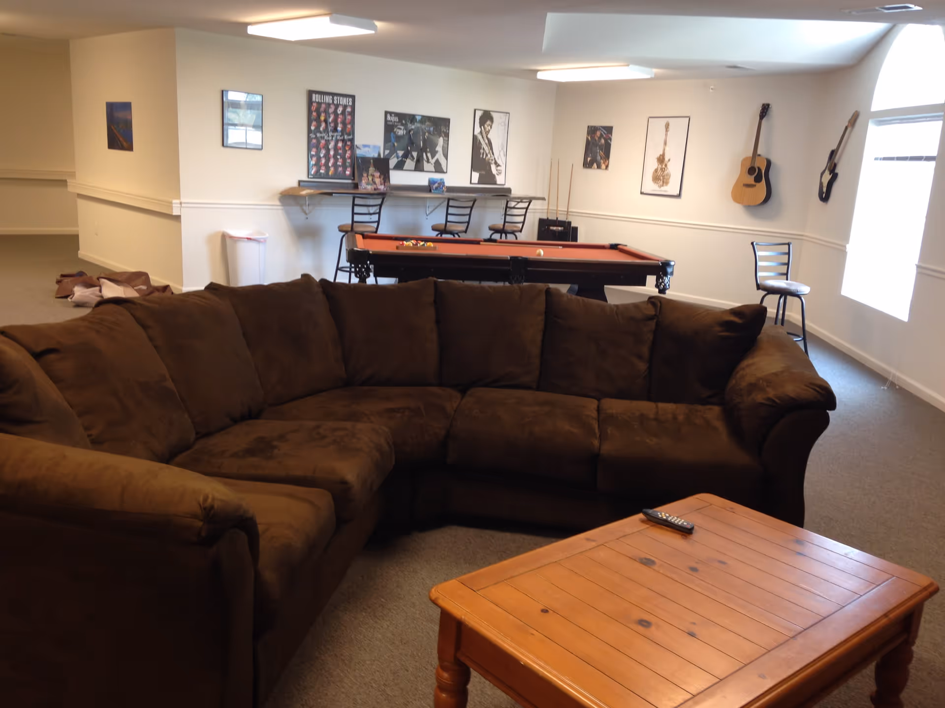 A cozy common area with a large brown sectional sofa and a wooden coffee table with a remote control on it. In the background, there is a pool table with balls set up, three bar stools at a counter, and several framed music posters on the walls. Two guitars hang on the wall near a window letting in natural light.