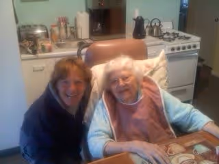 An elderly woman sitting at a table in a kitchen area with a younger woman beside her, both smiling. The kitchen includes a stove, kettle, and various kitchen items on the counter.
