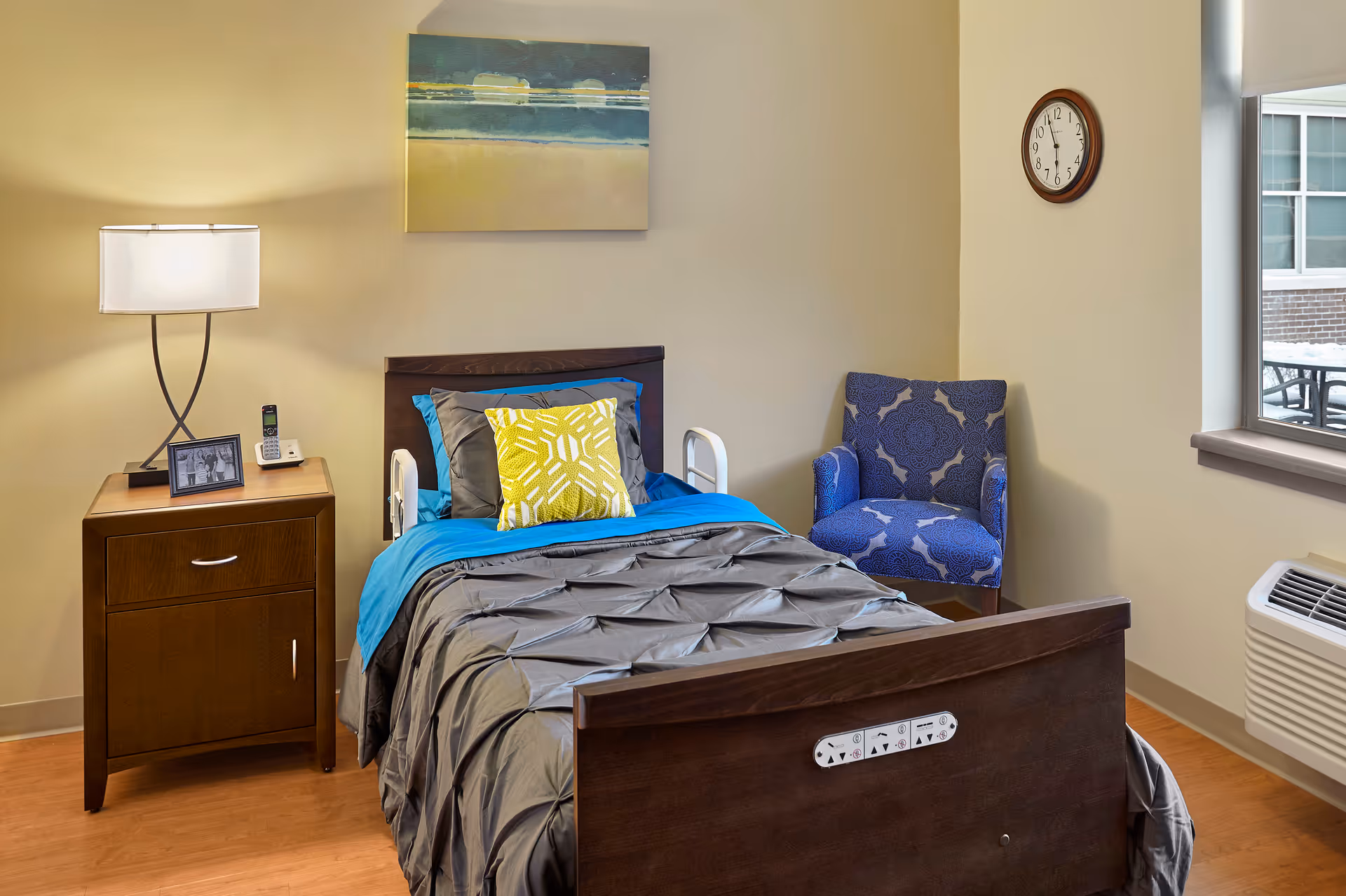 A neatly made single bed with a gray comforter, blue blanket, and yellow decorative pillow in a senior living facility room. Next to the bed is a wooden nightstand with a lamp, a framed photo, and a cordless phone. A blue patterned armchair is placed in the corner near a window with a view of an outdoor patio. A round wall clock shows the time as 12:05.