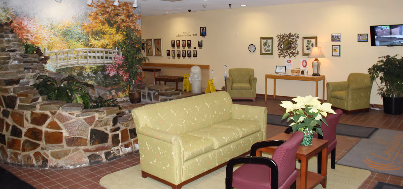 A cozy seating area in a senior living facility with a light green floral-patterned sofa, two purple chairs, and a wooden table with a white poinsettia plant. The background features a stone wall with plants and a mural of a white bridge over a stream surrounded by autumn trees. On the far wall, there are framed photos labeled 'Meet Our Administrative Staff,' a clock, decorative wall art, a lamp on a wooden table, and a TV screen. The floor is tiled with a rug near the seating area.