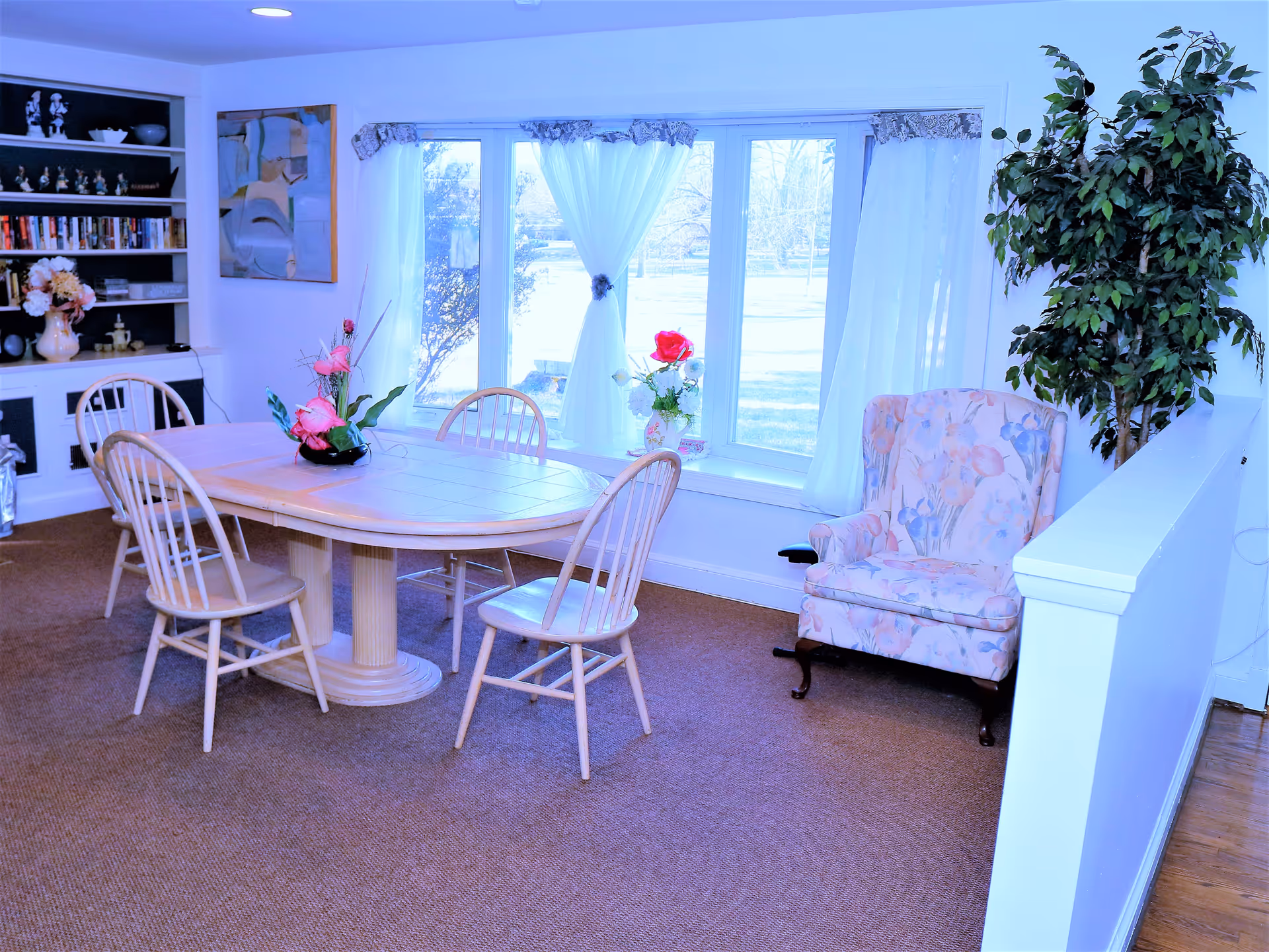 A bright dining area with a wooden table surrounded by four chairs. The table has a floral centerpiece with pink flowers. Behind the table is a large window with white curtains, letting in natural light. To the right of the table is a floral upholstered armchair next to a large green potted plant. On the left side, there is a built-in bookshelf filled with books and decorative items.